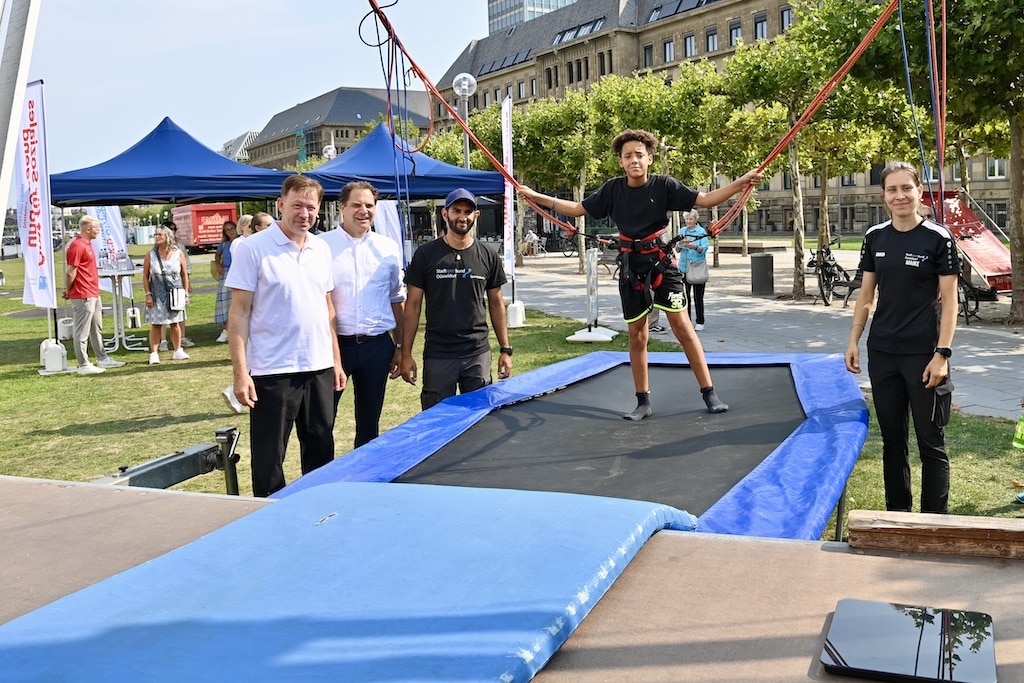 Startklar für das Olympic Adventure Camp 2025 auf der Rheinwiese am Apolloplatz: Stadtdirektor Burkhard Hintzsche (l.) und Rainer Klaren (2.v.l.) am Bungee Trampolin, auf dem Mehrfach-Saltos gemacht werden können. © Lokalbüro
