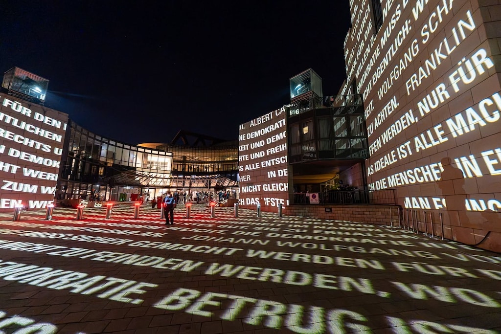 Auf Fassaden und Boden des Vorplatzes sind Sätze unter anderem von Albert Camus und Benjamin Franklin zu lesen. Im Hintergrund leuchtet der Eingang des Landtags. ©&nbsp;LB / Olaf Oidtmann
